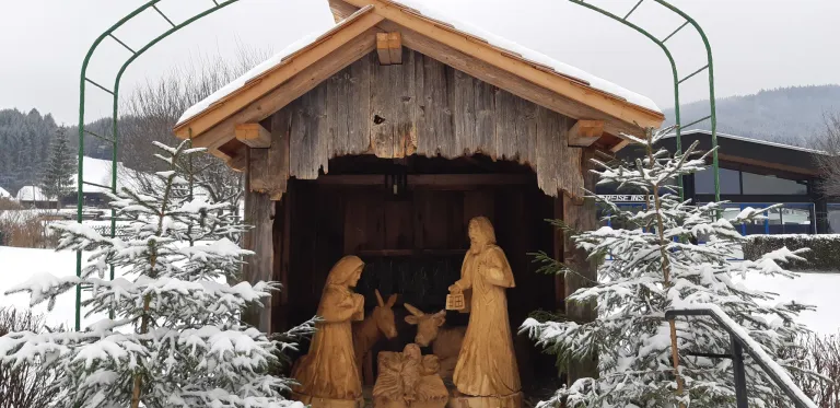 Eine hölzerne Krippe in einer rustikalen Scheune ist mit Schnee bedeckt. Sie zeigt Maria, Josef und das Jesuskind mit einem Esel und einem Schaf.