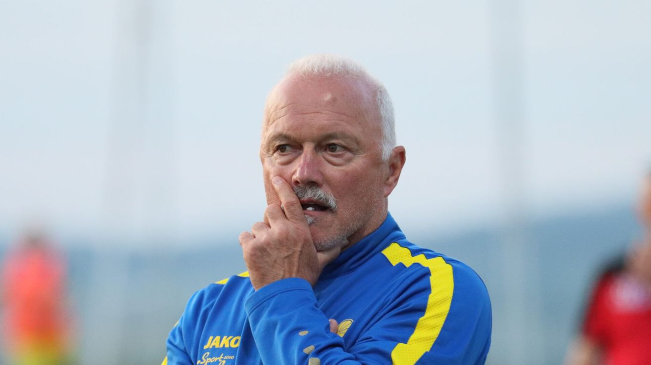 An older man with a mustache wearing a blue and yellow sports jacket with Jako and Sportcoo branding is looking to the left.