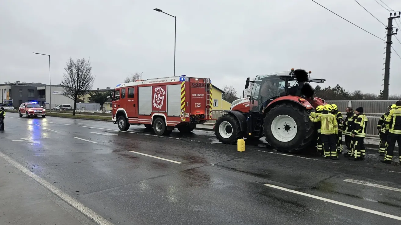 Ein roter Feuerwehrwagen ist neben einem Traktor auf einer nassen Straße geparkt. Beide Fahrzeuge haben Lichter an.