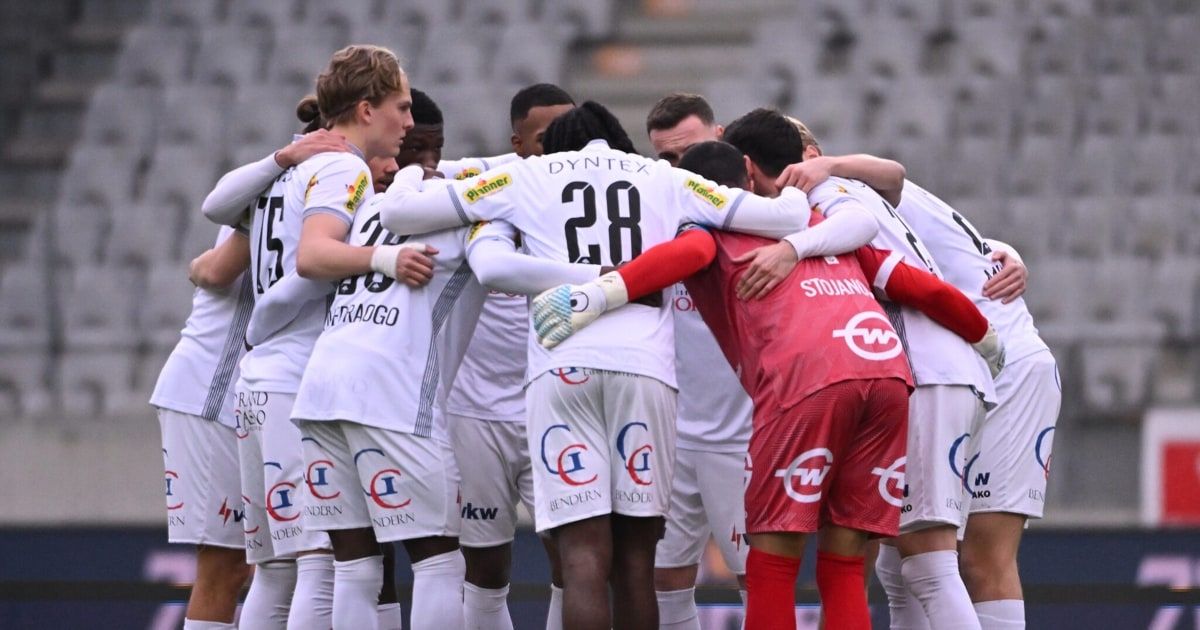 Soccer players in white jerseys huddle together on a field, with player number 28 in the center, while a player in a red jersey looks on.