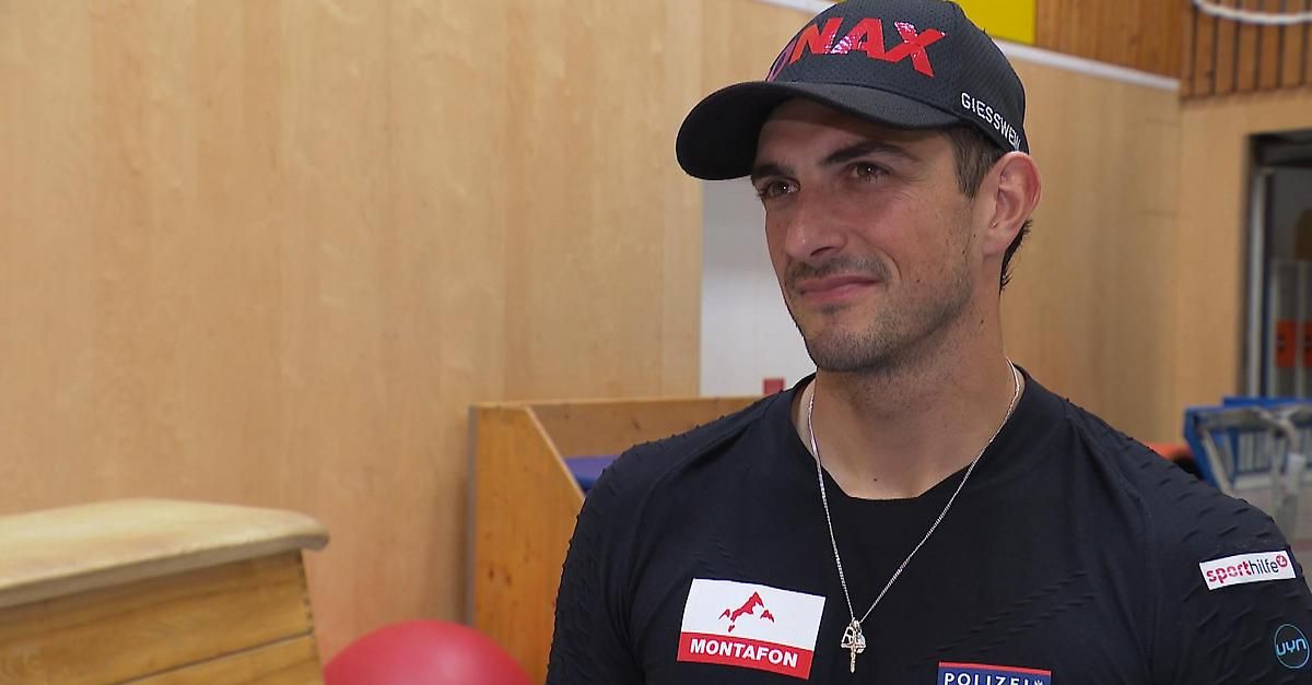 A man wearing a black cap with red text and a black shirt with a white tag is smiling. A red ball is in the corner of the room.