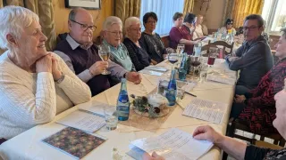 A group of older people are sitting around a dining table, holding wine glasses. They seem to be enjoying a meal. The table is set with plates, glasses, and napkins. There is a menu on the table. The room is dimly lit.