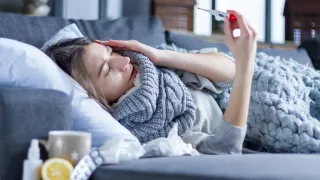 A woman lies on a couch, looking distressed, holding a thermometer, with tissues and a cup nearby.