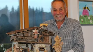 An older man smiles while holding a miniature nativity scene in front of a window.