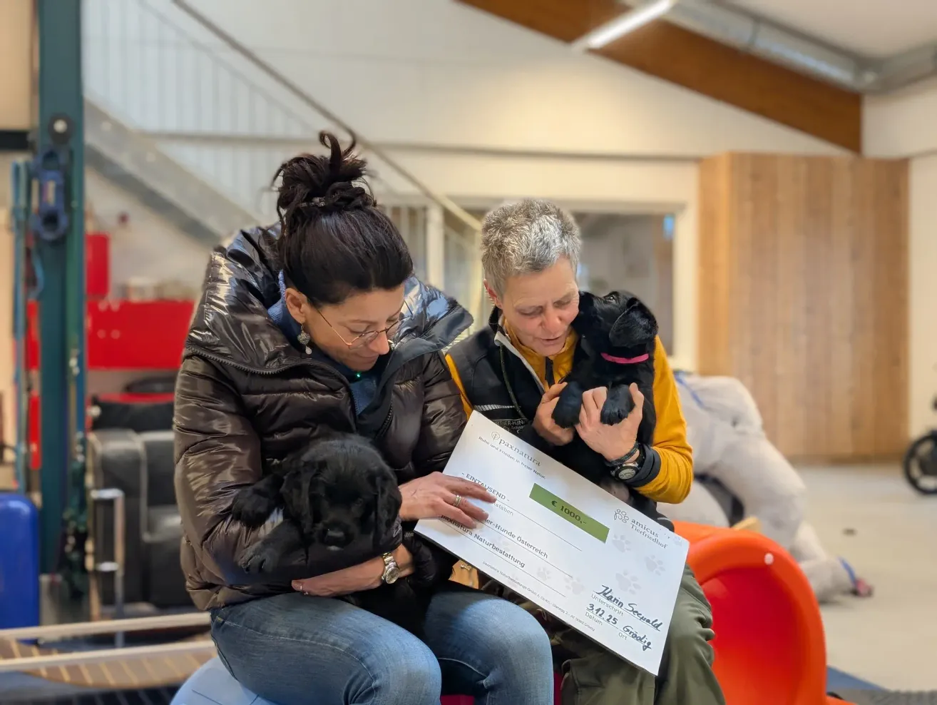 Two women sit on a cushion, holding black puppies and looking at a check with a large amount of money.