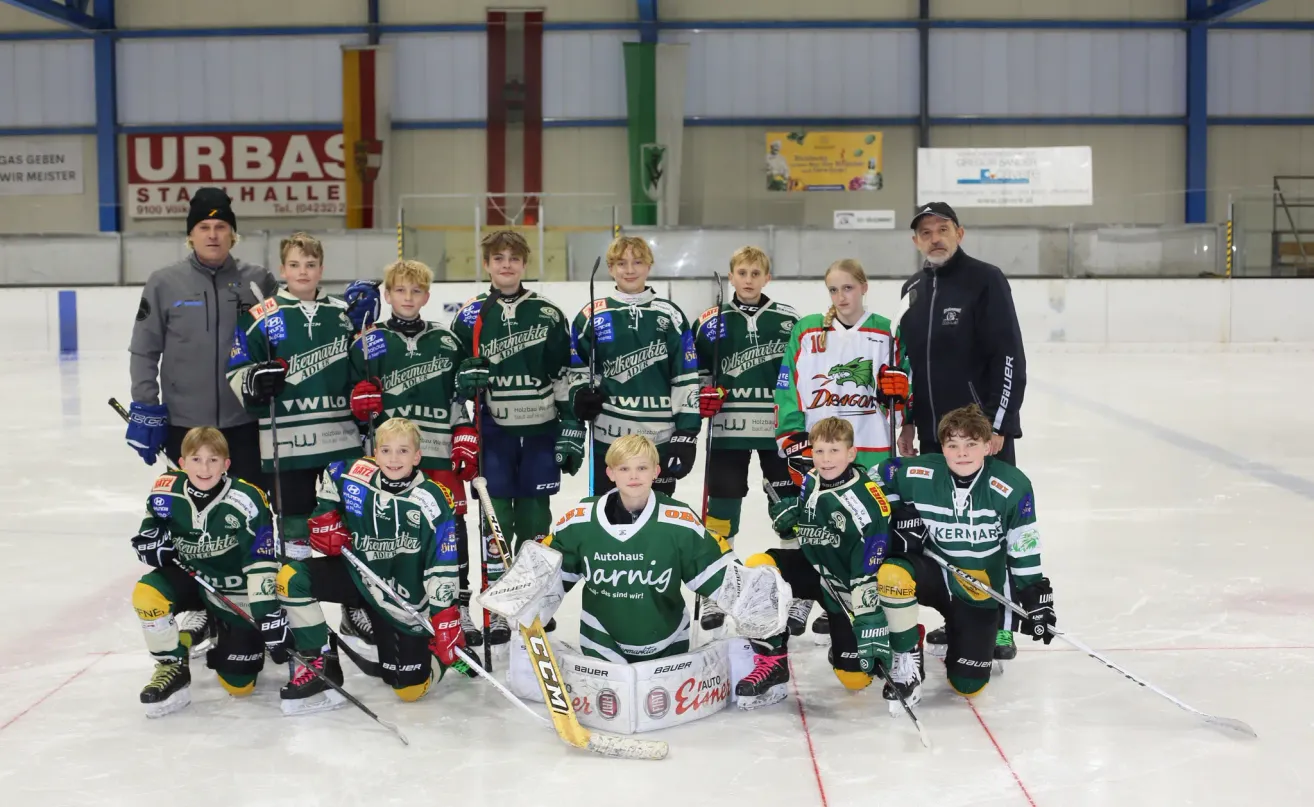 A youth hockey team poses for a photo in an ice rink, with players wearing green and white jerseys. An adult man stands behind them, wearing a black jacket. Banners and signs are visible in the background.