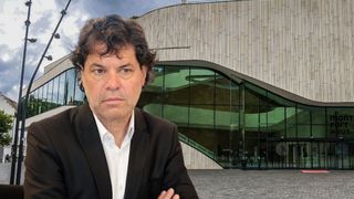 A man stands in front of a modern building, with his arms crossed. The building has a curved glass facade and a wooden wall.