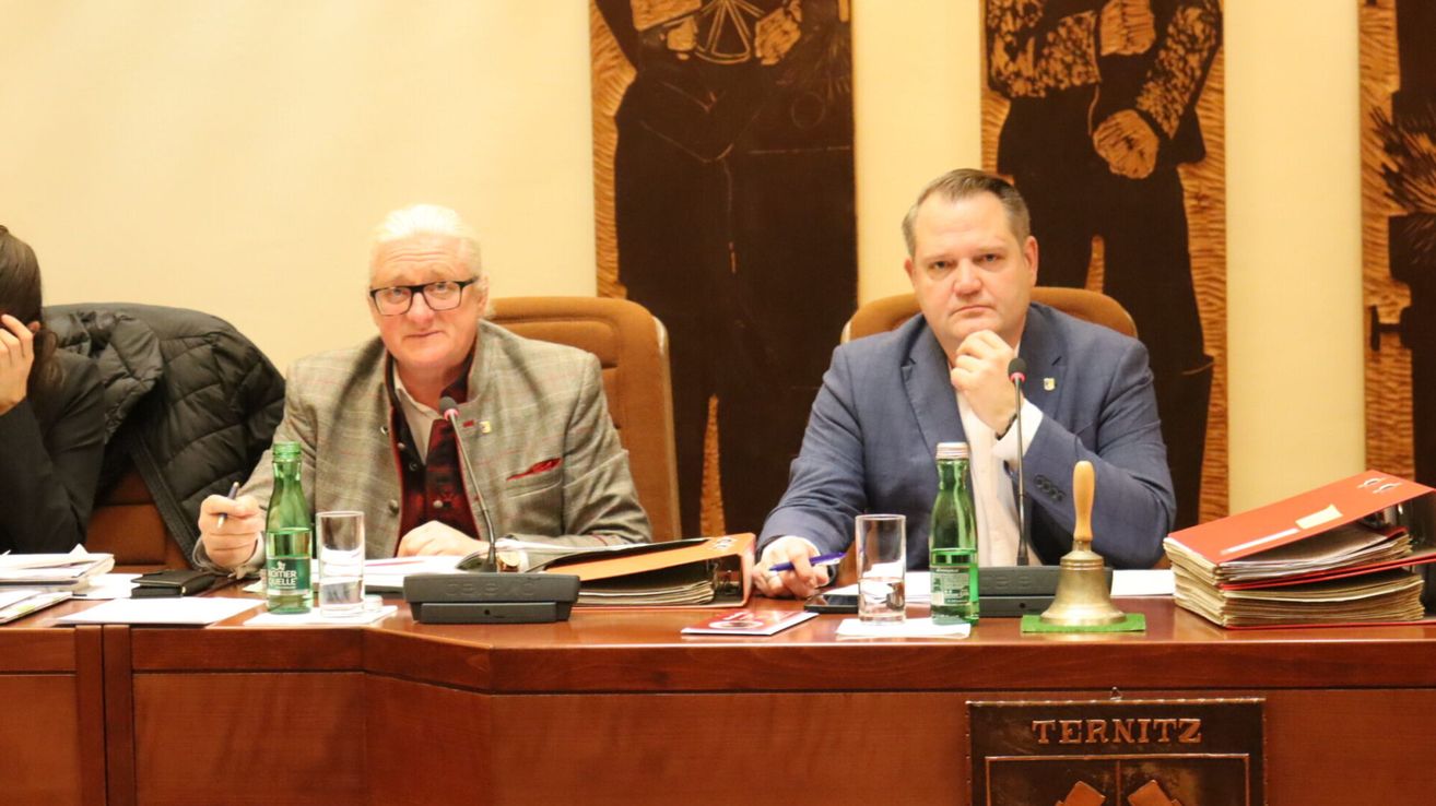 Two men are seated at a conference table, each with a microphone. The man on the left wears glasses and a blazer, while the man on the right has a serious expression. In front of them are a bell, a glass, and a green bottle.