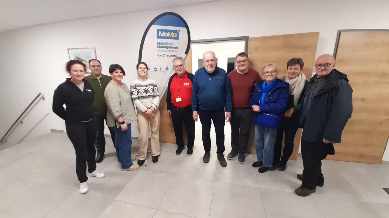 Eine Gruppe von Menschen steht vor einem Schild, auf dem 'Mobilitätsmanagement Niederosterreich noe@regional' steht. Sie lächeln und scheinen für ein Foto zu posieren.
