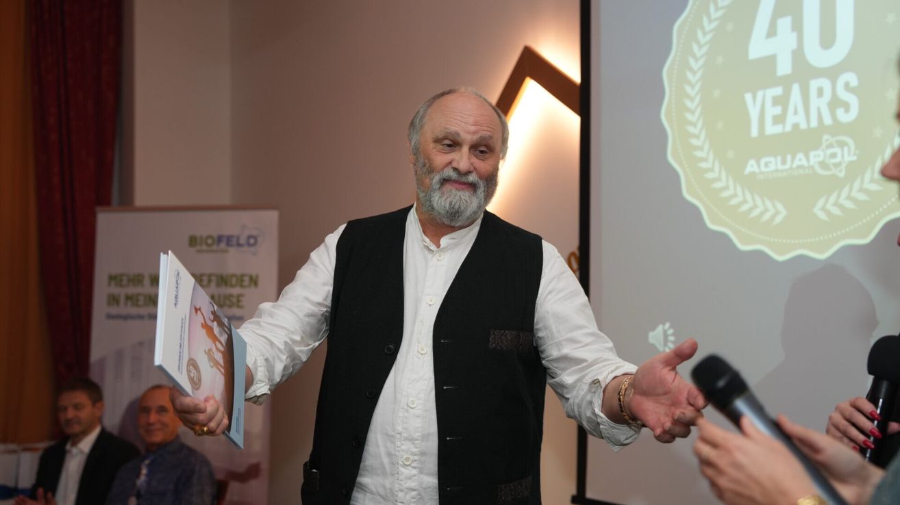 Ein älterer Mann in einem schwarzen Weste und weißem Hemd steht vor einer Projektionsleinwand und gestikuliert mit den Händen, während er ein Buch hält.