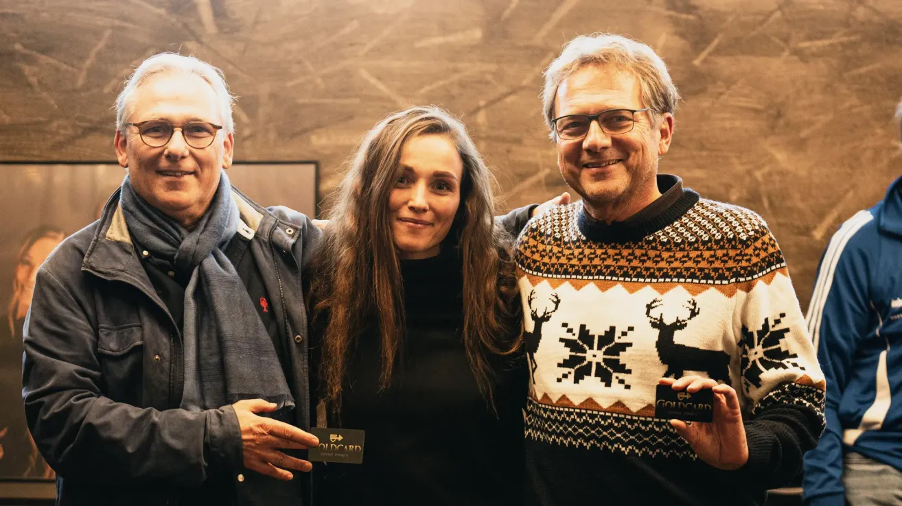 Drei Personen, zwei Männer und eine Frau, lächeln für ein Foto. Der Mann auf der linken Seite trägt eine Jacke mit einem Schal, während der Mann auf der rechten Seite einen Pullover mit einem Hirschmuster trägt. Die Frau trägt ein schwarzes Oberteil.