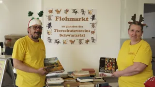 Two men in yellow shirts are standing in front of a table filled with books. They are smiling and appear to be at a book fair. A sign on the wall behind them reads Flohmarkt and Tierschutzvereins Schwarzwald.