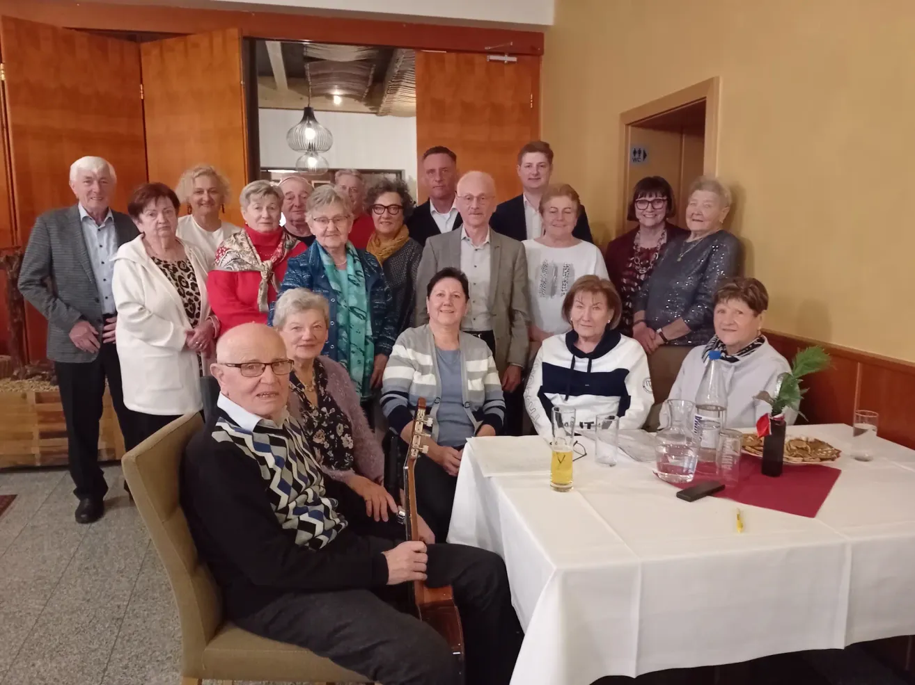 A group of elderly people, possibly a family, are gathered in a dining area for a family gathering. They are all smiling and seem to be posing for a photo.