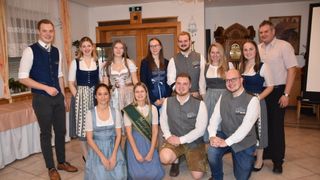 A group of individuals are posing for a photograph. Some are wearing traditional German attire. The two men in the front are kneeling. The woman in the middle is wearing a sash. Behind them is a wooden door and a clock.
