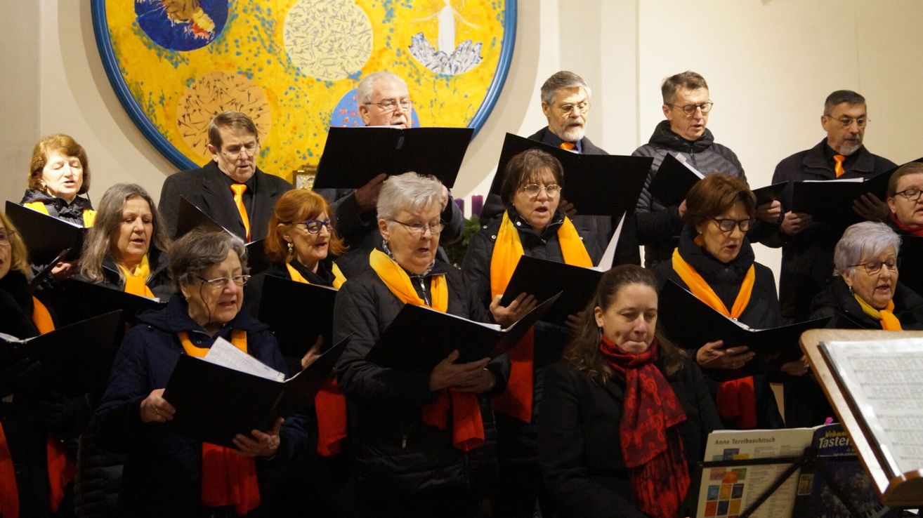 Ein Chor von Erwachsenen, einige mit Brillen, singen in einer Kirche, einige tragen Schals und halten Musikbücher.
