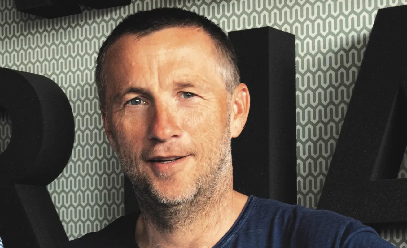 Ein Mann mit Bart und Schnurrbart lächelt in die Kamera. Er trägt ein blaues T-Shirt und steht vor einer geometrischen Tapete.