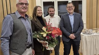 Four people standing in a room, smiling, and posing for a photo. A woman in the center holds a bouquet of flowers and a red gift box. Behind them, a glass door leads to another room.