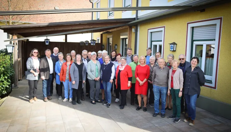 Eine Gruppe Erwachsener steht vor einem gelben Gebäude und lächelt für ein Gruppenfoto. Sie tragen verschiedene Kleidungsstücke und einige tragen Brillen. Das Gebäude hat einen Holzzaun und eine Überdachung.