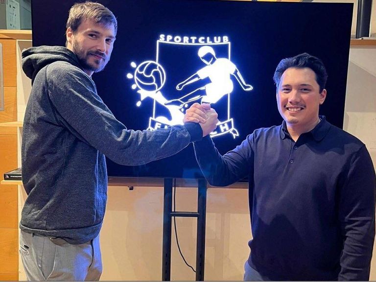 Two men smiling and shaking hands in front of a screen displaying a sports club logo.