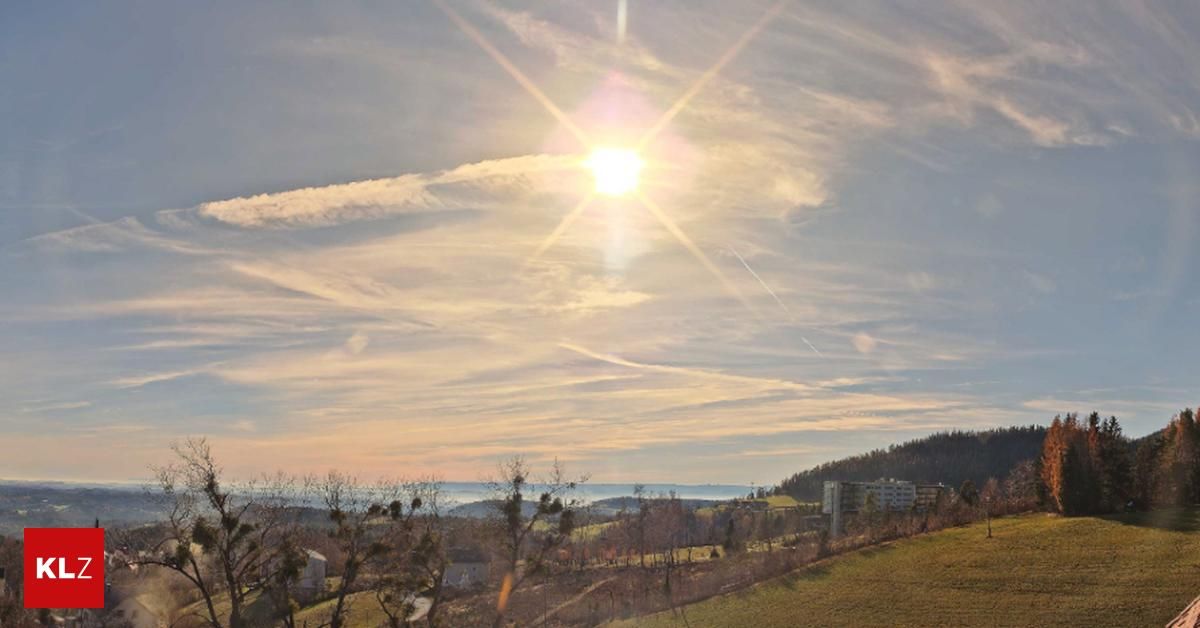 Eine strahlende Sonne scheint durch eine teilweise bewölkte Himmelslandschaft und wirft ein warmes Licht auf eine hügelige Landschaft. Bäume und Gebäude sind unten sichtbar.