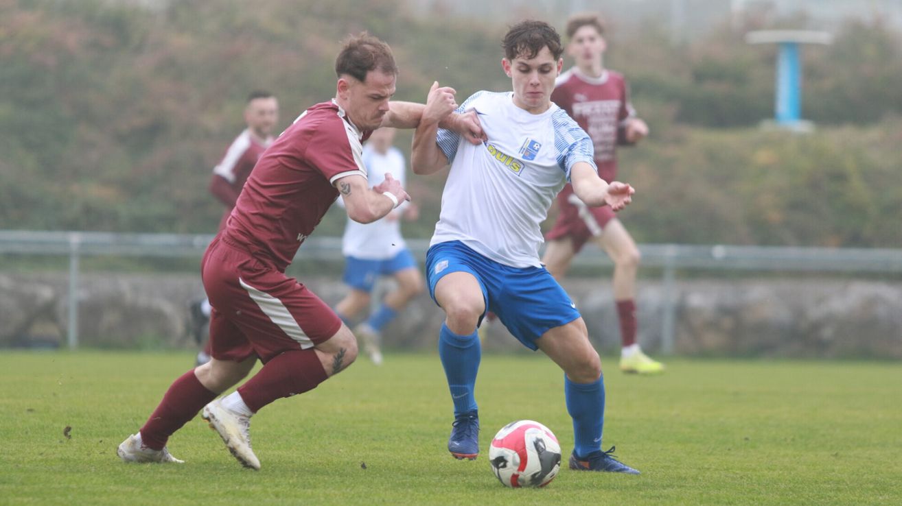 Zwei Fußballspieler kämpfen auf einem Feld um den Ball. Ein Spieler in einem weißen Shirt mit blauen Shorts sieht fokussiert aus.
