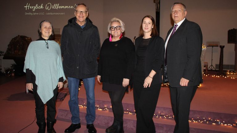 Fünf Personen stehen in einer Reihe auf einer Bühne, wahrscheinlich für ein Foto. Hinter ihnen ist eine Wand mit einem Kreuz und Text. Sie sind in formeller Kleidung gekleidet, wobei zwei Frauen Schwarz tragen. Die Bühne hat einen Hintergrund mit Lichtern.