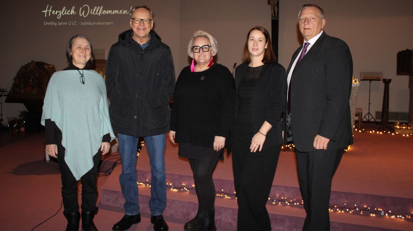 Fünf Personen stehen in einer Reihe auf einer Bühne, wahrscheinlich für ein Foto. Hinter ihnen ist eine Wand mit einem Kreuz und Text. Sie sind in formeller Kleidung gekleidet, wobei zwei Frauen Schwarz tragen. Die Bühne hat einen Hintergrund mit Lichtern.