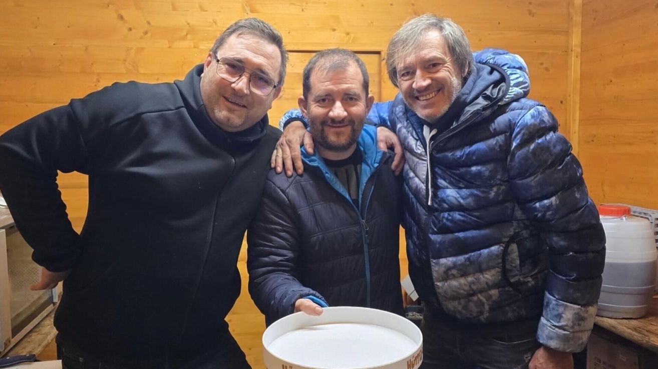 Drei Männer stehen nebeneinander, lächelnd, vor einem hölzernen Hintergrund. Einer hält eine weiße Schüssel.
