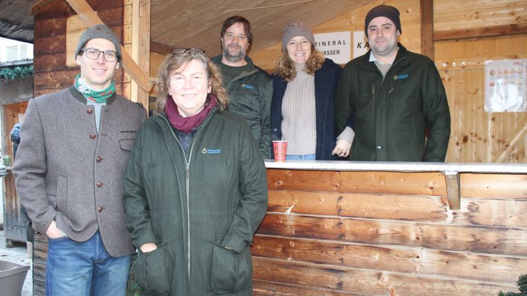Five people stand together in a wooden cabin. The woman in front has a scarf around her neck. Behind her, there is a counter with a cup on it. On the wall, there are signs with text.
