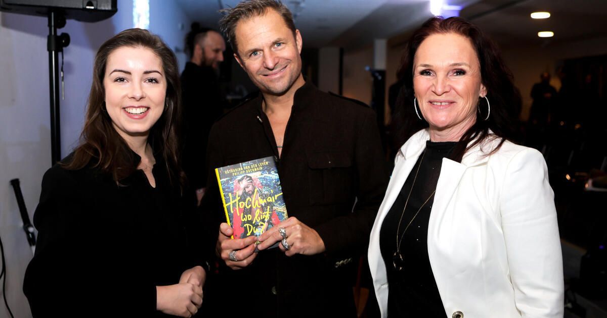 Drei Menschen stehen bei einer Veranstaltung eng beieinander und lächeln. Der Mann in der Mitte hält ein Buch mit dem Titel 'Hochzeit ohne Hut'.