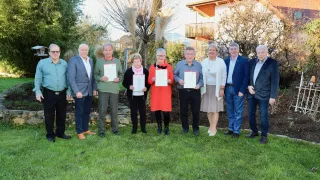 A group of individuals, possibly family, stand together on a lawn, smiling and holding certificates. Behind them, a house with a balcony is visible.