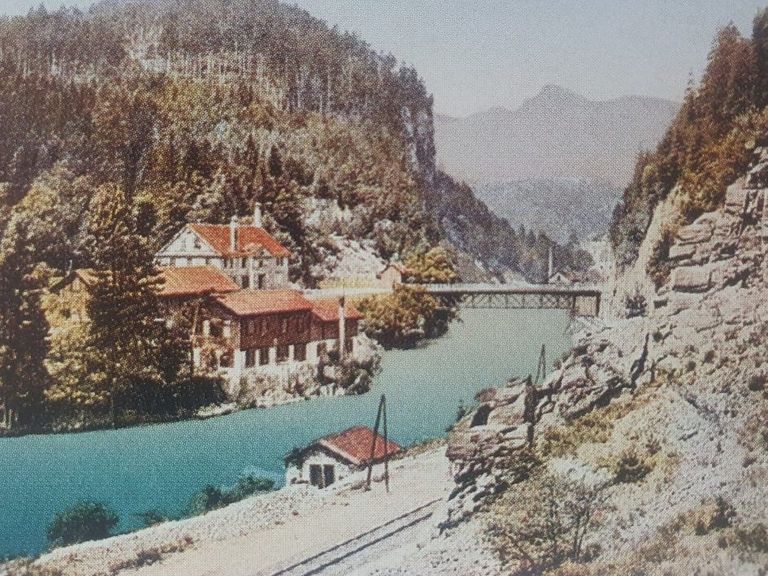 Ein historisches Foto einer bergigen Landschaft mit einem Fluss, einer Brücke und Gebäuden am Flussufer. Bäume umgeben das Gebiet.
