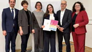 Five individuals, including a young girl holding a certificate, stand in front of a white banner with the word 'Architekt' on it. Behind them is a wall with the word 'Bildung'.