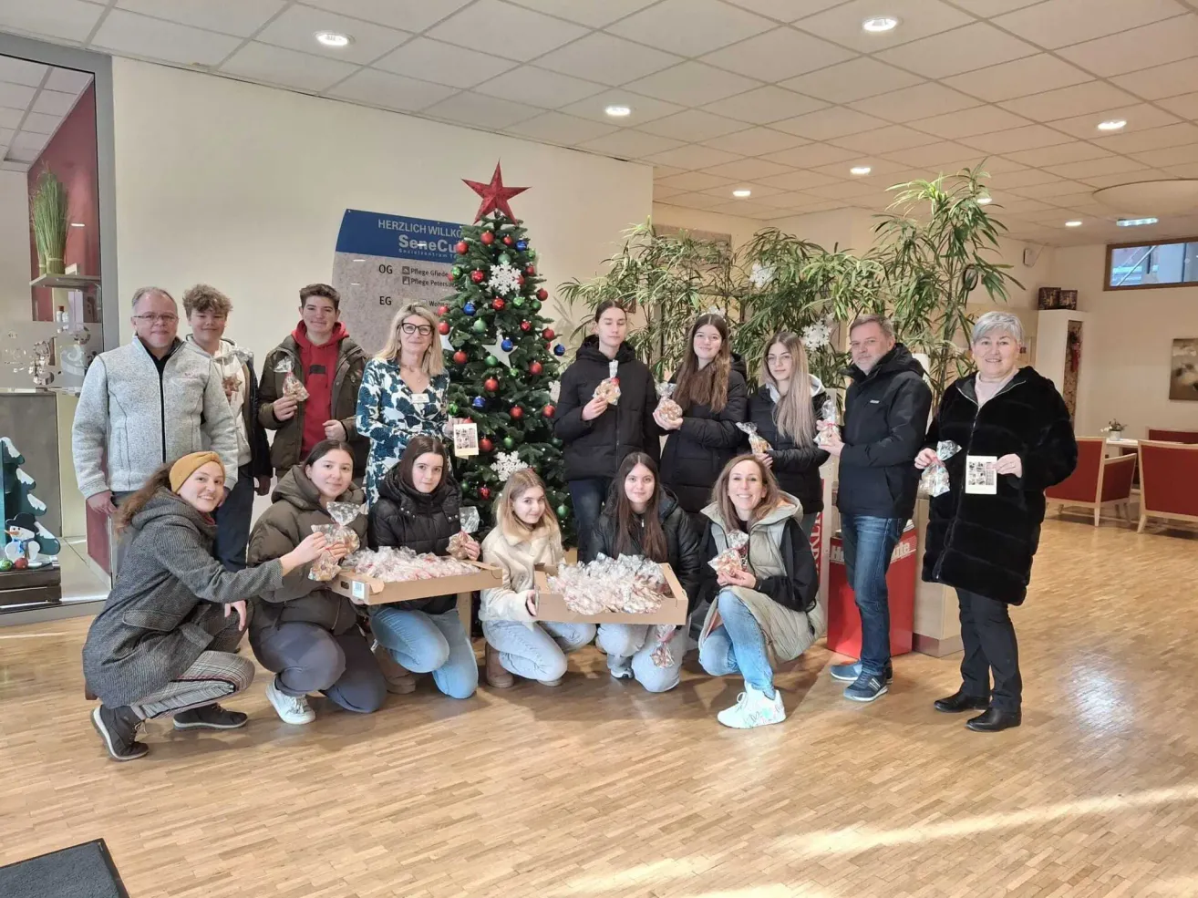 A group of people in winter clothing pose for a photo in front of a Christmas tree with gifts in their hands.