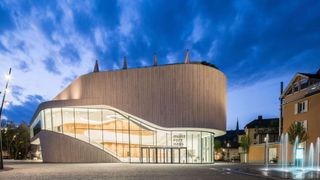 A modern building with wooden facades and glass windows. It has a curved roof with pointed spires. The building has a sign that reads 'Montfort Haus'.