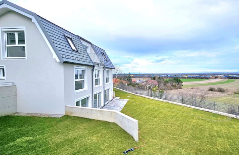 Ein modernes Haus mit einem grauen Dach und weißen Wänden. Es hat mehrere Fenster und einen kleinen Balkon. Das Haus ist von einem grünen Rasen umgeben. In der Ferne gibt es ein Feld mit einigen Bäumen und Häusern. Der Himmel ist bewölkt.