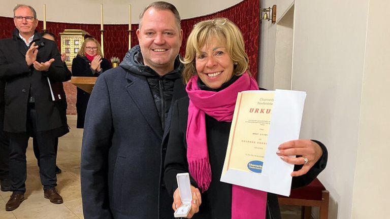 A man and a woman are smiling while holding a certificate and a ring box. The woman has a pink scarf. They are standing in front of a red curtain.