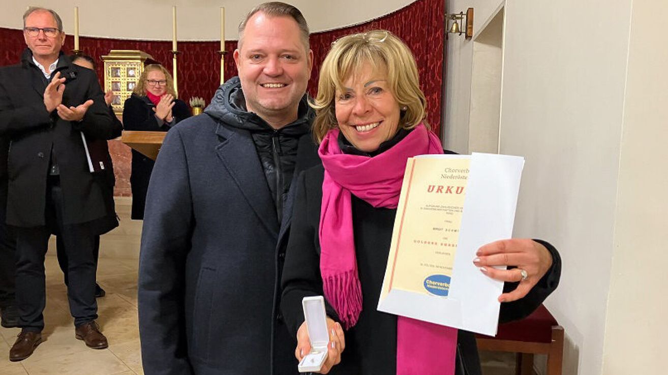 A man and a woman are smiling while holding a certificate and a ring box. The woman has a pink scarf. They are standing in front of a red curtain.