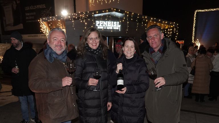 Four adults, two men and two women, stand outside a restaurant. They are smiling and holding wine glasses. The man on the left wears a scarf and a brown jacket. The woman on the right holds a bottle of wine. The restaurant has a sign that reads 'Peneist Born 2 Make Wine'.