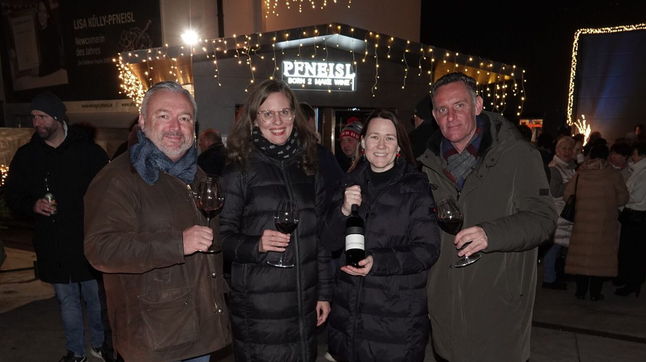 Four adults, two men and two women, stand outside a restaurant. They are smiling and holding wine glasses. The man on the left wears a scarf and a brown jacket. The woman on the right holds a bottle of wine. The restaurant has a sign that reads 'Peneist Born 2 Make Wine'.