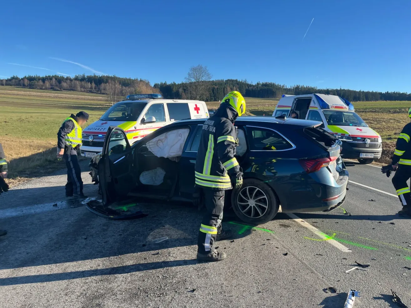 Emergency responders stand beside a damaged car, one of whom wears a yellow helmet. Behind, two ambulances are parked. The scene is on a road with green markings.
