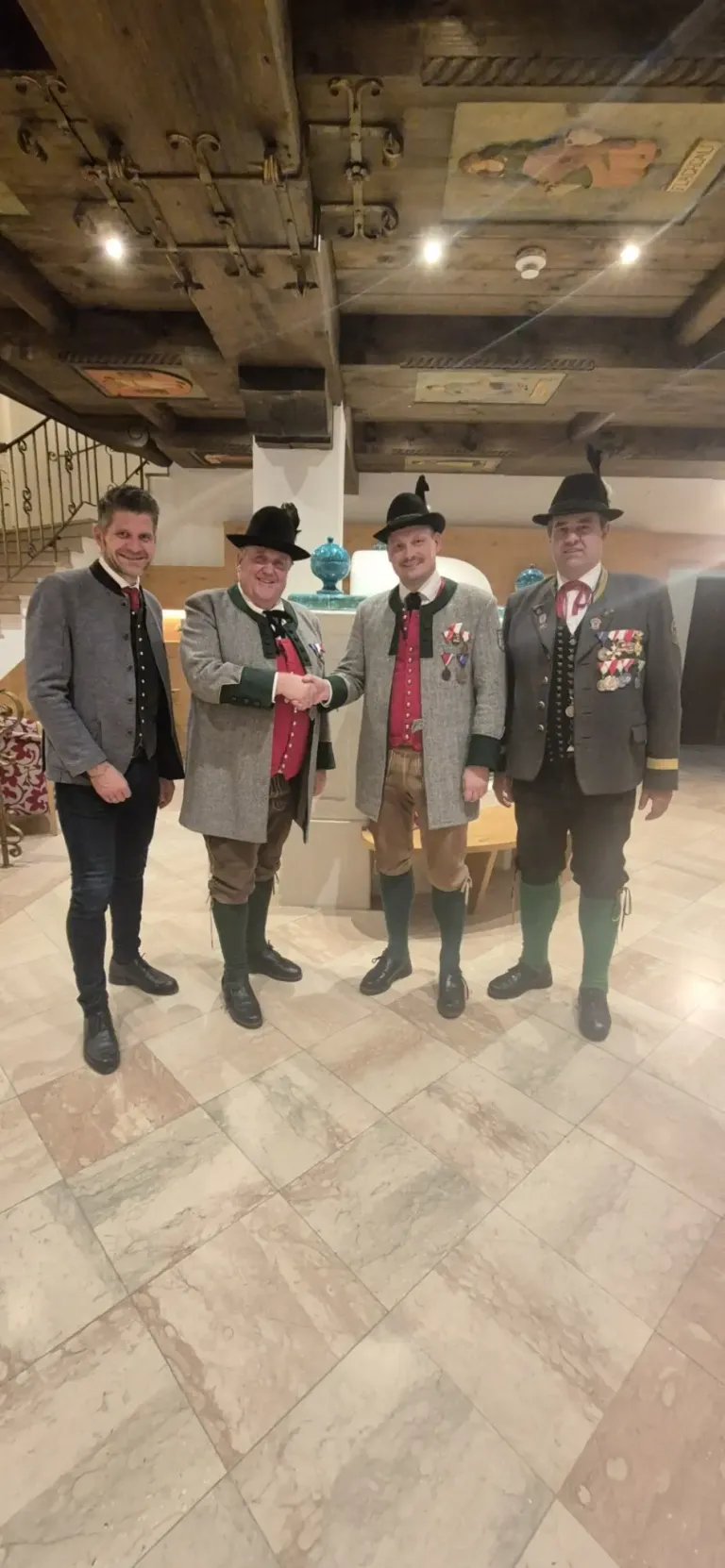 Vier Männer in traditioneller Kleidung stehen zusammen, einer davon schüttelt Hände. Sie tragen Hüte und Medaillen, und im Hintergrund befinden sich eine Treppe und eine Holzbank.