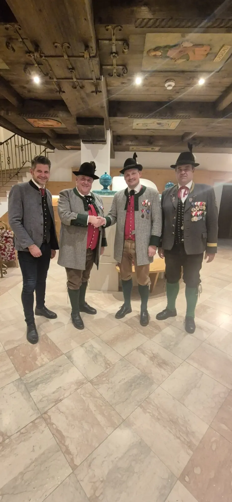 Vier Männer in traditioneller Kleidung stehen zusammen, einer davon schüttelt Hände. Sie tragen Hüte und Medaillen, und im Hintergrund befinden sich eine Treppe und eine Holzbank.