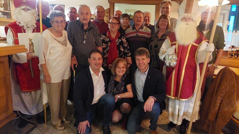 Eine Gruppe von Menschen, darunter ältere Personen und einige in formeller Kleidung, posiert für ein Foto mit dem Weihnachtsmann in einer festlichen Umgebung.