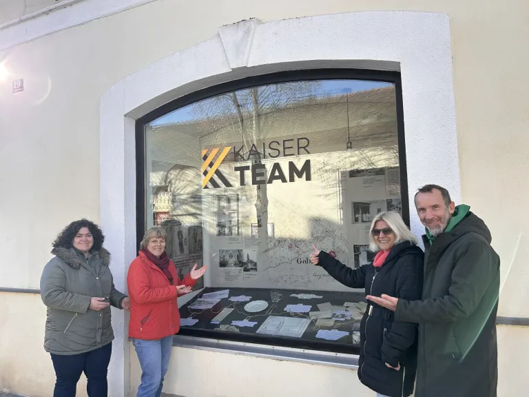 Vier Personen stehen lächelnd vor einer Kaiser Team Schaufenster-Ausstellung. Eine Karte und verschiedene Dokumente sind auf der Ausstellung.