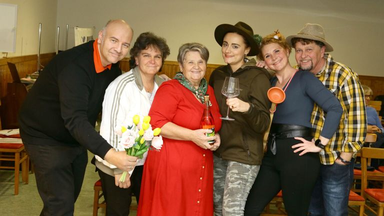 Eine Gruppe von Menschen posiert für ein Foto. Eine Frau hält Blumen, eine andere einen Weinglas und eine Frau in Rot hält eine Flasche. Die anderen tragen Freizeitkleidung.