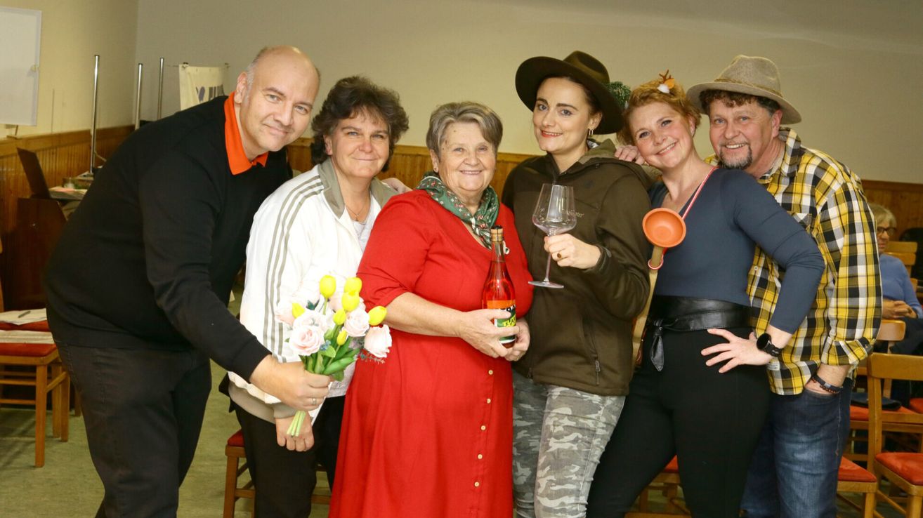 Eine Gruppe von Menschen posiert für ein Foto. Eine Frau hält Blumen, eine andere einen Weinglas und eine Frau in Rot hält eine Flasche. Die anderen tragen Freizeitkleidung.