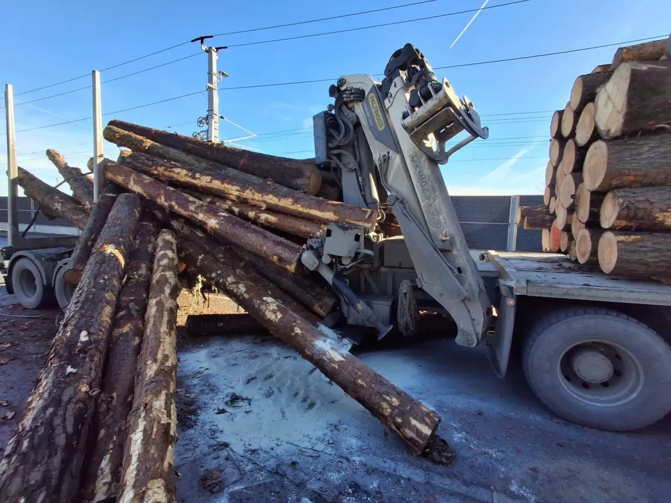 A crane is unloading logs from a truck on a road. The logs are piled up, and some are on the ground. The crane is on the right side.