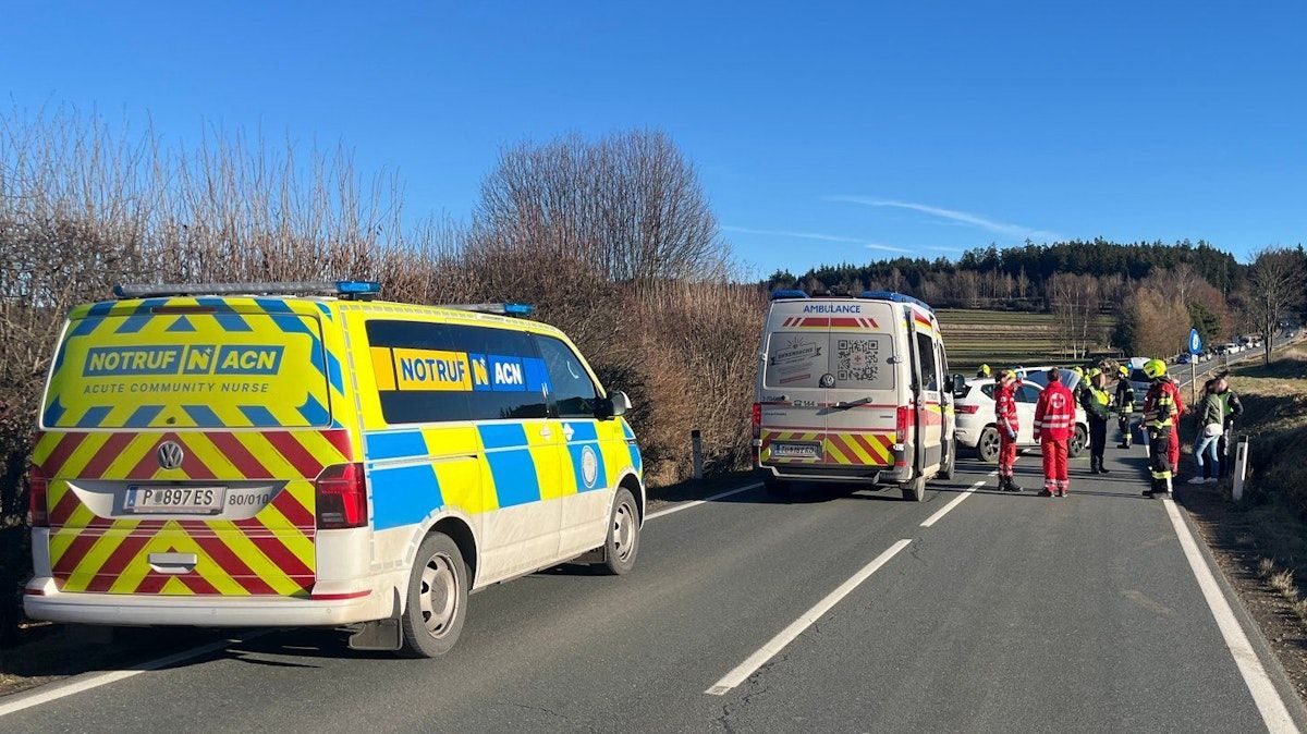 An ambulance and an emergency vehicle are parked on the side of a road. Two people in red uniforms are standing nearby.