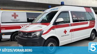 Two ambulances with red crosses on their sides, labeled 'Wiener Rotes Kreuz,' are parked in front of a building. The background shows a logo with 'Götzis/Vorarlberg' and a number '5'.
